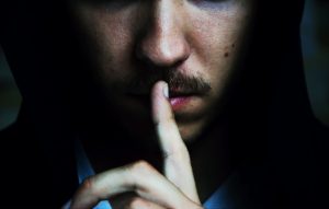 A man in a black hoodie with his finger on his lips, emphasizing the idea that silence is a sound.